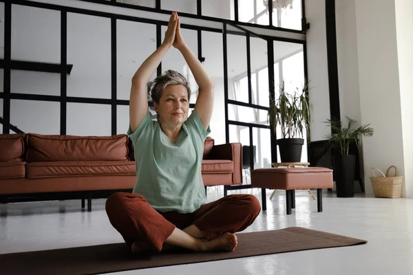 A middle aged lady practices yoga in the large hall of the house. A smiling woman sits on a mat on the floor in the lotus position and shows namaste gesture with palms, raised above head. Looks at the