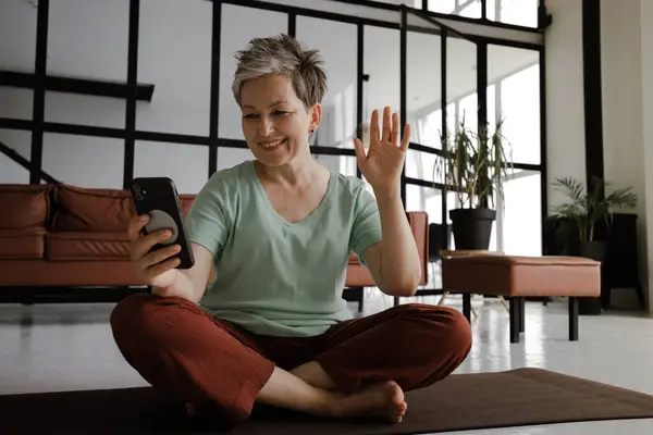A middle aged lady practices yoga in the large hall of the house. A smiling woman sits on a mat on the floor in the lotus position, has a break time from training, uses a smartphone for video