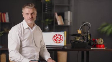 Ciddi bir orta yaşlı grafik tasarımcısı masanın kenarında oturur. İçinde geometrik katının üç boyutlu kırmızı modelinin çizimi olan bir laptop. Bir 3D yazıcı makinesi otomatik olarak çalışır