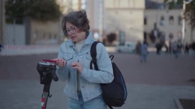 Güzel yaşlı kadın şehirde elektrikli scooter 'a binmek için akıllı telefon kullanıyor. Kadın elektrikli scooter 'ın parasını ödemek ve kilidini açmak için telefon uygulaması kullanıyor. Yüksek kalite 4k görüntü