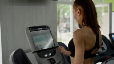 Kendine güvenen genç bir kadın spor salonunda antrenman yapıyor. Koşu bandı ayarları monitöründeki bir düğmeye basar ve adımlar atmaya başlar. Yoğun çalışmadan önce ideal ısınma. Yüksek kalite 4k görüntü