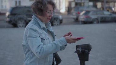 Güzel yaşlı kadın şehirde elektrikli scooter 'a binmek için akıllı telefon kullanıyor. Kadın elektrikli scooter 'ın parasını ödemek ve kilidini açmak için telefon uygulaması kullanıyor. Yüksek kalite 4k görüntü
