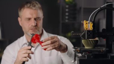 Orta yaşlı bir grafik tasarımcısı dijital kalibreler kullanır ve kırmızı plastik huninin parametrelerini ölçer. Otomatik özerk süreçte çalışan ve haki rengi üreten 3D yazıcı makinesi