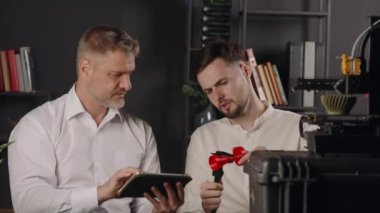 Genç bir makine mühendisi dijital kalibreler kullanır ve kırmızı plastik huninin parametrelerini ölçer. Orta yaşlı bir grafik tasarımcısı sonuçları tablete kaydeder. 3D yazıcı haki rengi yapar
