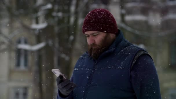 一个留着胡子的成年人在户外用智能手机戴着红帽子 男人在收到送信人的迷因时面带微笑 在寒冷的天气聊天 下雪时 带着胡子的丈夫在社交媒体上冲浪 — 图库视频影像