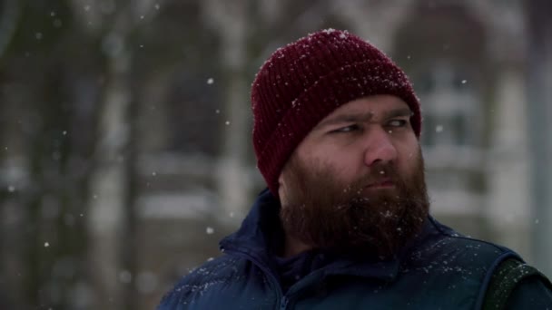 雪が降ったときにひげを被った深刻な残忍な男のスローム 屋外で雪を降りながら待っている大人のひげをした男 赤い帽子とジャケットを着ている男性は 冬を嫌っているようです — ストック動画