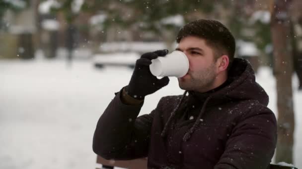 ベンチに座ってコーヒーを飲んで 美しい雪が降るのを見ている若者をスロー クリスマスの時間の概念 屋外の魔法 男性は外で温かいお茶を飲み 雪が降っている間に夢を見ます 高品質で — ストック動画