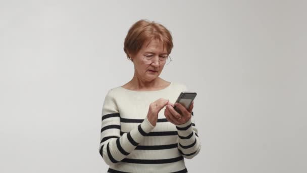 一位老年妇女戴着眼镜 手持并使用智能手机打字并回复留言 一位年长的 心满意足的微笑女士优雅地点击屏幕 在积极向上的情绪中筛选答案 — 图库视频影像