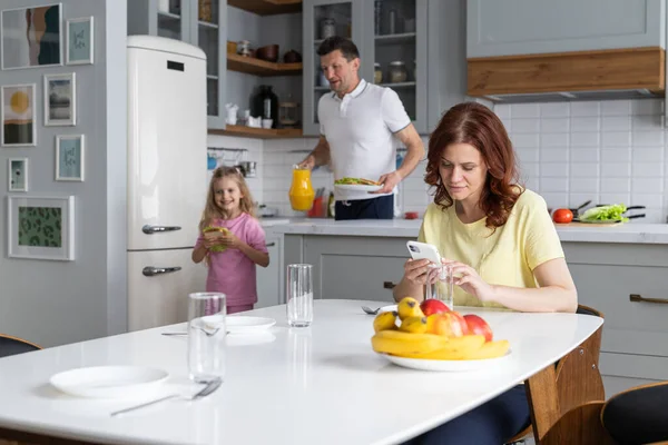 An american dream friendly family in the kitchen in a joyful mood prepare for lunch together. A young man father and smiling daughter set the table, while mother sits and rests with smartphone. The