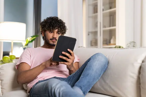 A smiling mestizo guy reclining on the sofa in the room, enjoys relax after work or study in the cozy home environment. A positive young african or american man holds a tablet in hands and watch a