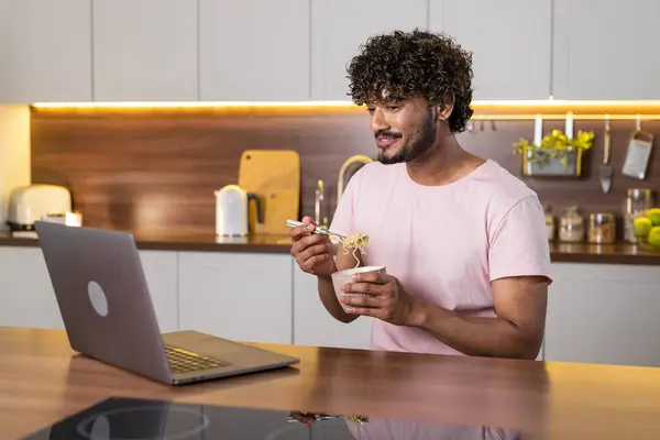 A mestizo young man stands sideways in the kitchen in subdued lighting, holds a bowl of pasta in hands and wraps them in a fork. An american or african guy student uses a laptop, enjoys a dinner and
