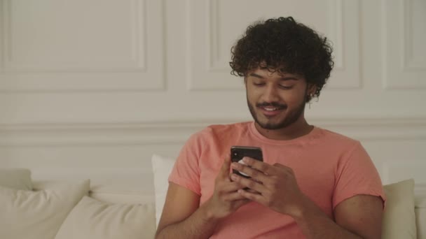 魅力的でポジティブなアフリカ系アメリカ人の学生は 自宅のリビングルームのソファーに座り 勉強した後に休む 若い笑顔の男が手を握り スマートフォンを面白く見ている — ストック動画