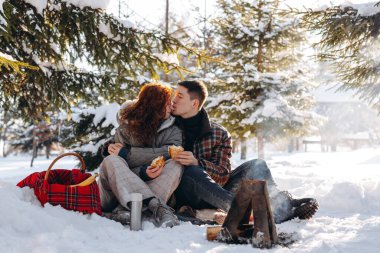 Evli bir çift karlı bir ormanda piknik yaptı. Genç insanlar küçük bir ateşin önünde oturup krep yerler. Yüksek kalite fotoğraf
