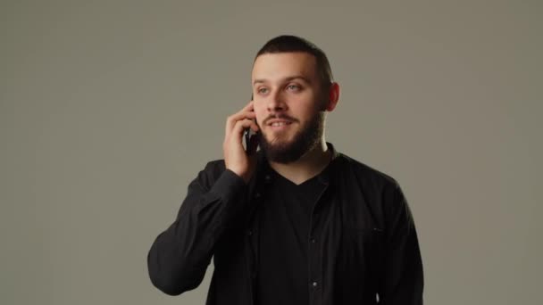 黒いシャツを着た若い髭の男が電話で話していて 灰色の背景に孤立している 高品質4K映像 — ストック動画