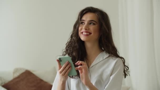 笑顔のブルネットがベッドの上に座って 彼女のスマートフォンで何かを見ています 快適な若い女性は彼女の電話で彼女のソーシャルネットワークを閲覧しています 高品質4K映像 — ストック動画
