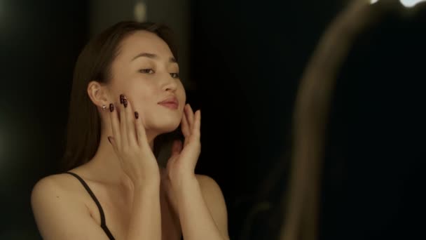 鏡の前に立つ若いアジア系の女性は 頬とあごを指で優しく撫でている 美しいブルネットが鏡の前で彼女の顔を調べる 高品質4K映像 — ストック動画
