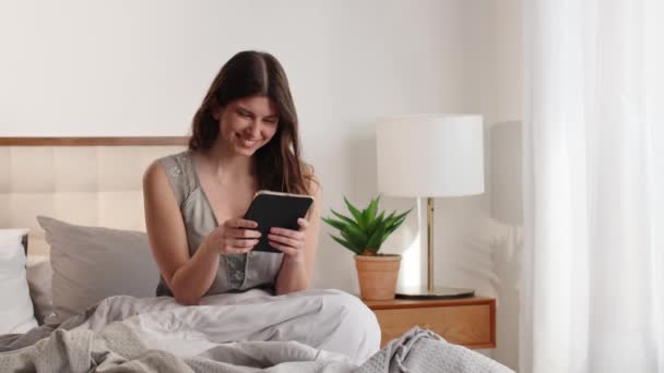 Uma Jovem Morena Usa Tablet Digital Enquanto Senta Uma Cama — Vídeo de Stock
