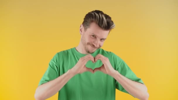 陽気若い男は手で心臓の兆候を示しています 黄色の壁に屈強なポーズで笑顔の若い男は 心臓の兆候を示しています 高品質4K映像 — ストック動画