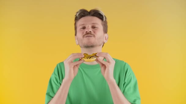 頑固な青年がケーキの味を楽しんでいる 緑のTシャツの男が黄色い背景のカメラの前に立ってケーキを食べる 高品質4K映像 — ストック動画