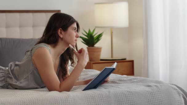 かわいいブルネットがベッドの上に横たわり ノートブックにメモを作ります 巻き毛の若い女性がベッドの上に横たわっており 週末のためのTo Doリストを作ります 高品質のフルHd映像 — ストック動画