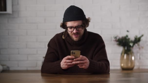 穿着棕色毛衣头戴灰色帽子的男人发短信给他的女朋友 当他的男朋友收到女朋友用智能手机拍的照片时 他很高兴 整天发短信 在厨房用智能手机的人高 — 图库视频影像