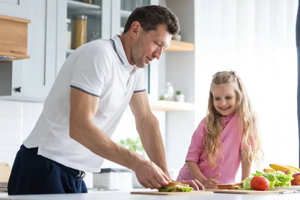 一个父亲和女儿友好的家庭站在厨房里管理着 一位严肃的年轻人爸爸教他的孩子独立做三明治 相互关系的概念 图库照片