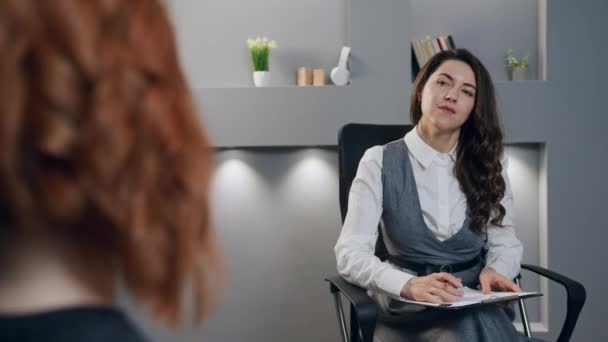 若い女性心理学者女性は椅子に座り 赤毛患者からの情報を記録しています 精神科医または神経科医との相談 若い女性は心配して個人的療法中にノート — ストック動画