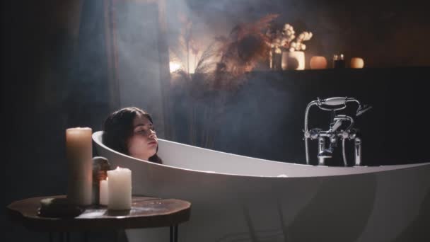 一个女人正在享受一个轻松的浴池 背后闪烁着一支蜡烛 她的下巴轻轻地放在大腿上 嘴角挂着柔和的微笑 — 图库视频影像