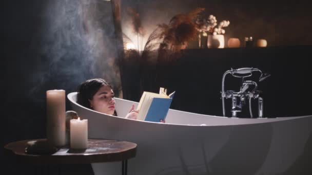 一个女人正在享受一个轻松的浴池 背后闪烁着一支蜡烛 她的下巴轻轻地放在大腿上 嘴角挂着柔和的微笑 — 图库视频影像