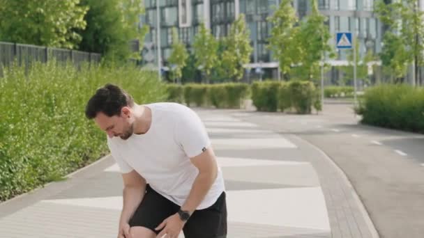 一个年轻的运动健将停止慢跑 腿和脚受伤 扭伤或脱臼 一个运动员坐在人行道上 痛苦地面带皱纹 站起身来抱着膝盖 — 图库视频影像