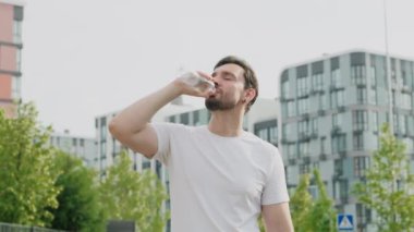 Pozitif ruh hali içinde genç bir sporcu antrenmandan sonra dinlenir, alnını siler ve şehir parkındaki susuzluğunu giderir modern konut binaları arasında. İlham verici bir gülümseme