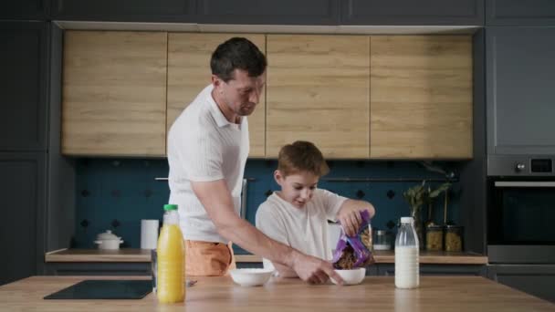 一个由父子组成的快乐的家庭准备在厨房里吃早饭 一个有爱心的男孩帮助爸爸 把饼干从包里撒在碗里 一个男人尝到了甜酸苦辣的滋味 对待孩子 — 图库视频影像