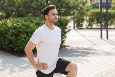 Genç bir adam parktaki bir kasabada yanlamasına çömeliyor. Sporcu bir adam koşmadan önce bacak kaslarını ısıtır. Sağlıklı bir yaşam tarzı olarak fiziksel eğitim kavramı. Yüksek kalite fotoğraf