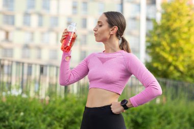 Sıska genç bir kadın elinde bir şişe tutuyor, şehir caddesinde yan duruyor, modern konut binalarıyla çevrili. Kendine güvenen ve memnun sportif bir kız antrenmandan sonra dinlenir. Bir