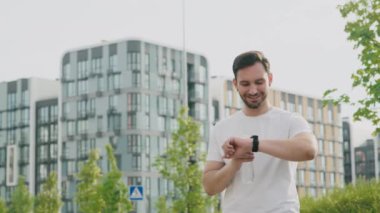 Genç sportif bir adam şehir parkının dışında duruyor, saati kontrol ediyor, modern yüksek binalar arasında keyifli bir toplantı bekliyor. İlham verici gülümseyen bir sporcu.