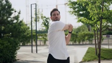 Genç bir sporcu sabah egzersizi yapar, şehir parkında koşmadan önce kollarını ve boynunu uzatır. Bir atlet, park alanında antrenman ya da yarışma için hazırlanıyor.