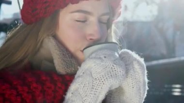 Örgü eldivenli genç, hayalperest kadın sıcak çay içiyor. Termostan kahve fincanı. Kış kenti parkında sıcak içkinin tadını çıkaran tatlı bir kız arkadaş. Gülümseyen ve sıcak içeceğin tadını alan genç bir kadın. Yüksek kalite 4k