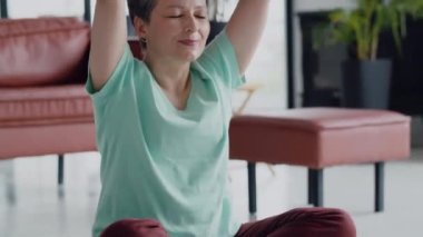 Yaşlılıkta hoş ve kullanışlı bir eğlence anlayışı. Yaşlı kadın evde yoga egzersizi yapıyor. Olgun kadınlar meditasyon yapar. Kameraya bakar ve gülümser. Yüksek kalite 4k görüntü