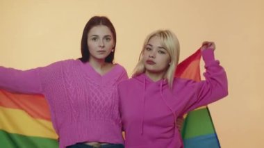 Bej renkli arka planda izole edilmiş gökkuşağı bayrağına sarılmış mutlu bir şekilde sarılan birkaç genç lezbiyen kız. Genç lezbiyenler, LGBT bayrağına sarılmış kadınları kucaklıyor. Oryantasyon toleransı