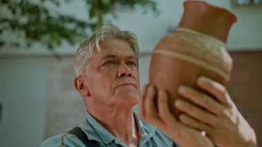 Çömlekçilikten anlayan yaşlı adam yeni bir çömlek kapını tutuyor. Profesyonel çömlekçi yeni sanat eserlerini açık havada seyrediyor. Yetişkin erkek el yapımı çiğ seramik gösteriyor.