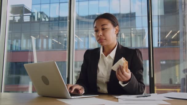 若いアフロ系アメリカ人の女性は 片手で何かをタイプし 別のものと一緒に食べます オフィスの黒人女性が朝食にサンドイッチを食べるのは時間がないからだ ストレスの多い仕事と不健康な食事 — ストック動画