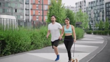 Genç ve sportif bir aile sabah evcil hayvanıyla koşuyor, Jack Russell Terrier köpeği, şehir parkında yüksek binaların arasında. Arkadaşlık ve arkadaşlık kavramı.