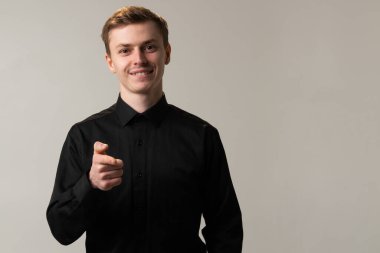 Resmi giyinmiş, siyah gömlekli ve ceketli bir adam yüzünde bir gülümsemeyle kameraya bakıyor. Kolunu, yakasını ve çene çizgisini gösteriyor..