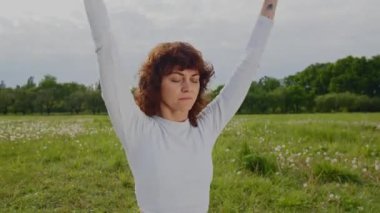 Beyaz eşofmanlı genç bir kadın yoga eğitiminde ellerini kaldırıyor. Sporcu kadın, Pranayama meditasyonu üzerine yoğunlaşmıştı. Kız parkta elini kaldırır ve zihnini açar. Yüksek kalite 4k görüntü
