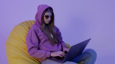 Pouf 'un üzerinde oturan ve mor renkli arka planda izole edilmiş not defteri kullanan genç bir kadın. Kapüşonlu kız uzaktan kumandalı çalışıyor ve puf minder üzerinde yatıyor. İnternette anonim olduğu için güneş gözlüklü komik kız. Hacker