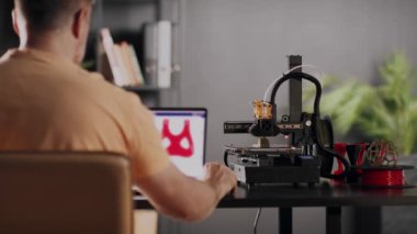 Bir erkek grafik tasarımcısı dizüstü bilgisayar ekranında tasvir edilen kalp ve göğüs için kırmızı renkte ameliyat sonrası sabitleme bandajı modeli geliştirir. Bir 3D yazıcı makinesi otomatik olarak çalışır