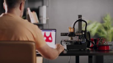 Bir erkek grafik tasarımcısı dizüstü bilgisayar ekranında tasvir edilen kalp ve göğüs için kırmızı renkte ameliyat sonrası sabitleme bandajı modeli geliştirir. Bir 3D yazıcı makinesi otomatik olarak çalışır