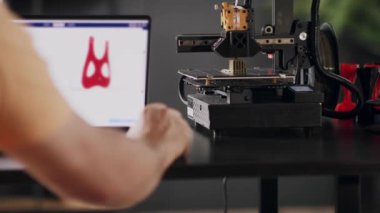 Bir erkek grafik tasarımcısı dizüstü bilgisayar ekranında tasvir edilen kalp ve göğüs için kırmızı renkte ameliyat sonrası sabitleme bandajı modeli geliştirir. Bir 3D yazıcı makinesi otomatik olarak çalışır