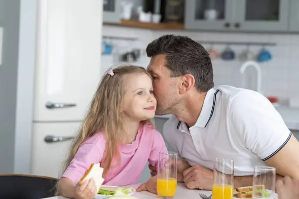 一个来自父亲和女儿友好的家庭坐在餐桌旁 一起在厨房里吃午饭 一个年轻人爸爸紧紧地搂着小女孩 在她耳边低声说着一个秘密 图库图片
