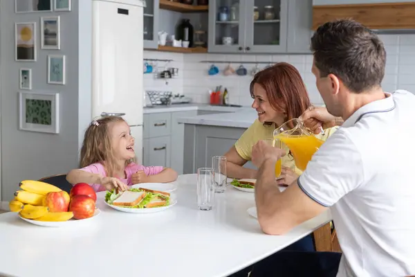 一个美国梦中的友好家庭 在厨房里欢快地准备着午餐 一位年轻男子的父亲和笑着的女儿摆好桌子 而母亲则坐着用智能手机休息 免版税图库图片